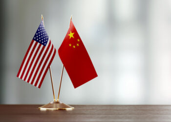 American and Chinese flag pair on desk over defocused background. Horizontal composition with copy space and selective focus.